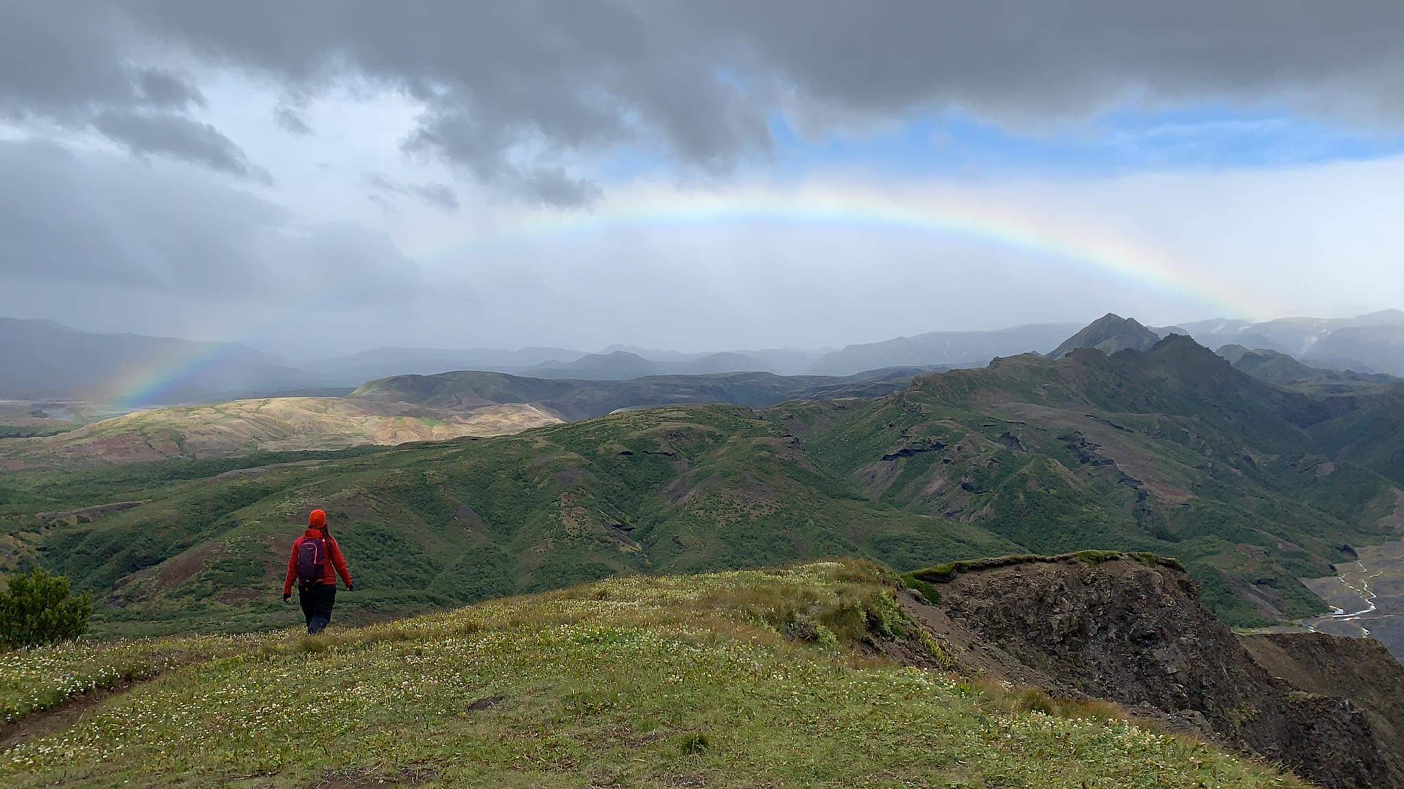 A hiker in a red jacket walking along a green mountain ridge beneath a rainbow